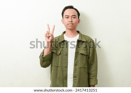 asian man posing smiling and looking at camera showing two (2) finger. Portrait of Indonesian man in green jacket on white background isolated.