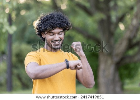 Close-up photo of a happy young Indian male athlete standing in a park and enjoying the result on his smartwatch, showing a trendy gesture of success and victory.