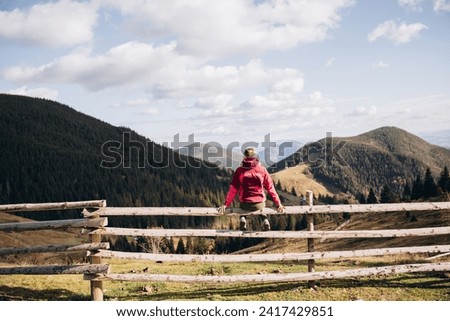  a woman in a pink ski jacket sits on a wooden fence overlooking the mountains. travel in the mountains Royalty-Free Stock Photo #2417429851