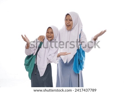 Two excited Indonesian student wearing hijab and uniform carrying bags standing isolated while rising hands