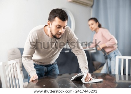Young family couple keeping their apartment clean. Bearded man wiping furniture with rag while his wife hoovering couch.. Royalty-Free Stock Photo #2417445291