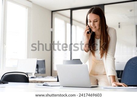 Young businesswoman in a dynamic work pose, speaking on the phone and using her laptop, which highlights her ability to handle multiple tasks efficiently in a fast-paced work environment Royalty-Free Stock Photo #2417469603