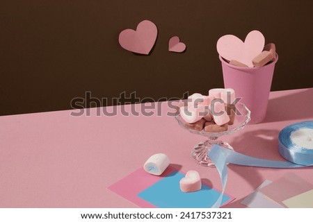 A stack of colorful papers decorated with a glass tray and pink cup of marshmallows. A roll of blue ribbon featured. Origami hearts pinned on brown background. Space for product presentation