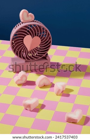 A cute purple handheld fan and heart-shaped marshmallows are displayed against a pink-yellow checkered background. Sweet colors for advertising.