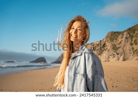 Portrait of smiling woman with long blond hair on sandy beach. Happy female in denim shirt with sea and mountain in background. Lady enjoying vacation against blue sky.