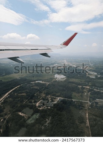 A view from an airplane window during flight, capturing part of the plane’s wing and the landscape below. The airplane’s wing is grey with a red-tipped winglet. Royalty-Free Stock Photo #2417567373