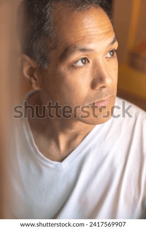 Vertical portrait of young Japanese man gazing out of a window with a pensive expression. Concept related to mental health, contemplation, introspection, or melancholy. Royalty-Free Stock Photo #2417569907