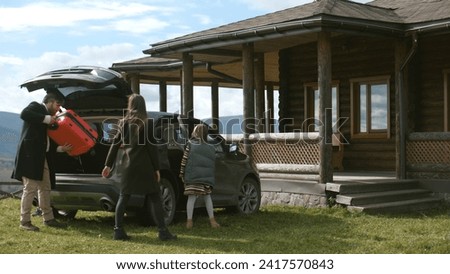 Happy family gets out of the car, walks with suitcase to modern house. Couple with kids settles into rented wooden cottage in mountains for vacation or moves to new home property. Real estate concept. Royalty-Free Stock Photo #2417570843