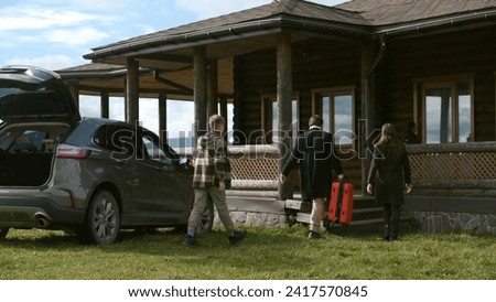 Happy family gets out of the car, walks with suitcase to modern house. Couple with kids settles into rented wooden cottage in mountains for vacation or moves to new home property. Real estate concept. Royalty-Free Stock Photo #2417570845