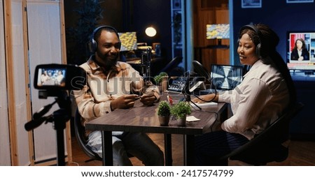 Man and woman laughing during comedy focused podcast filmed with professional camera. African american guest amused by talking show host telling jokes, recording with high tech equipment Royalty-Free Stock Photo #2417574799