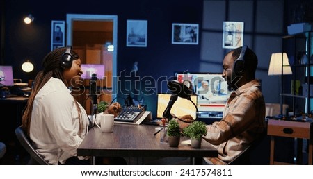 Vlogger show host adjusting analog mixer while talking with guest in neon lights ornate personal studio, ensuring perfect sound quality. Man and woman filming online podcast episode, zoom out shot Royalty-Free Stock Photo #2417574811