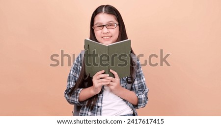 Cute school girl is carrying a school bag and holding a notebook. Isolated on brown background in studio. Royalty-Free Stock Photo #2417617415
