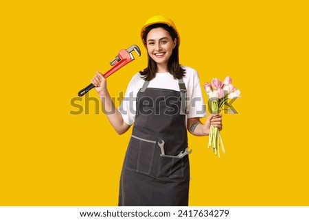 Portrait of young female construction worker with bouquet of flowers and pipe wrench on yellow background. International Women's Day Royalty-Free Stock Photo #2417634279
