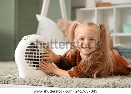 Cute little girl warming near electric fan heater in bedroom Royalty-Free Stock Photo #2417637843