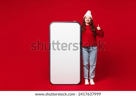 Full body young woman wearing knitted sweater white hat casual clothes big huge blank screen area mobile cell phone smartphone show thumb up isolated on plain red color background. Lifestyle concept