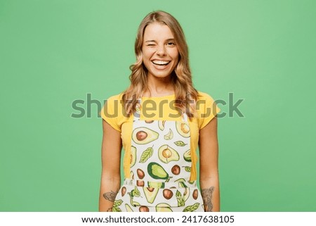 Young smiling happy satisfied creative housewife housekeeper chef cook baker woman wears apron yellow t-shirt look camera wink isolated on plain pastel green background studio. Cooking food concept