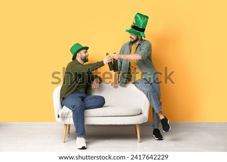 Young men in leprechaun hats with green beards holding bottles of beer and sitting on sofa near yellow wall. St. Patrick's Day celebration