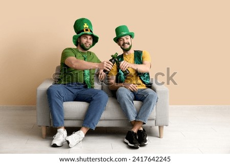 Young men in leprechaun hats with green beards holding bottles of beer and sitting on sofa near beige wall. St. Patrick's Day celebration
