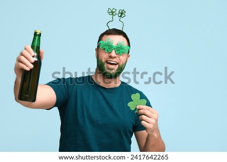 Young man in headband with green beard holding bottle of beer and clover on blue background. St. Patrick's Day celebration