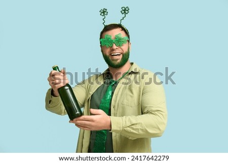 Young man in headband with green beard holding bottle of beer on blue background. St. Patrick's Day celebration