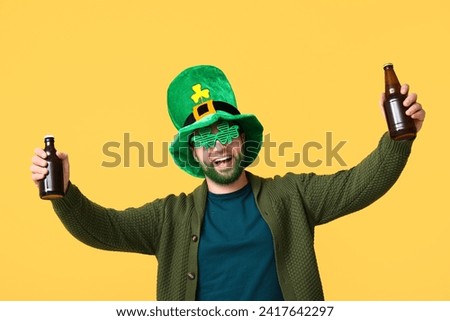 Happy young man in leprechaun hat and decorative glasses in shape of clover with green beard holding bottles of beer on yellow background. St. Patrick's Day celebration