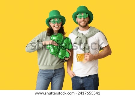 Beautiful young couple in leprechaun hats with air balloon in shape of clover and glass of beer on yellow background. St. Patrick's Day celebration