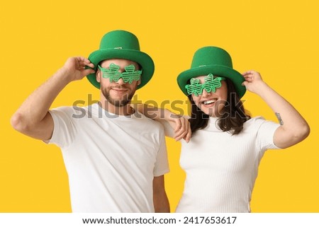 Beautiful young couple in leprechaun hats and decorative glasses in shape of clover on yellow background. St. Patrick's Day celebration Royalty-Free Stock Photo #2417653617