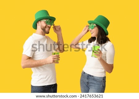 Happy young couple in leprechaun hats with glasses of beer on yellow background. St. Patrick's Day celebration