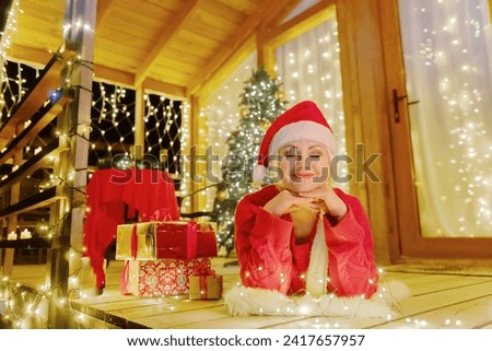Woman in Santa hat by Christmas tree. Festive setting. Illustrating holiday cheer and celebration. She is posing for a picture, and there are presents nearby, adding to the festive atmosphere.
