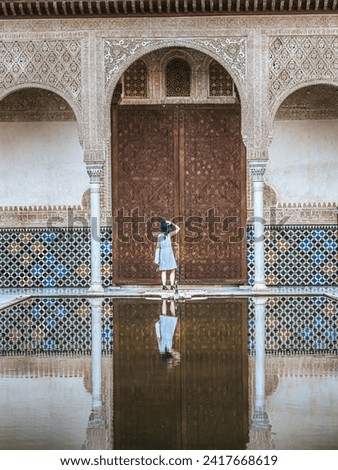 A beautiful young woman female tourist standing in front of the Alhambra door in a blue dress and hat, reflection in the water in the Courtyard of Myrtles, solo travelling, tourism, visitor, Granada