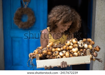 Smiling Woman Looking to her Homegrown Onions in the Crate she is Holding in front of Her Organic Farm House in the Countryside