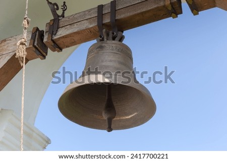 Bronze cup-shaped church bell hung to wooden pivoted beam called headstock to swing to and fro in the arched belfry on yard of Christian church, view against the clear sky 
 Royalty-Free Stock Photo #2417700221