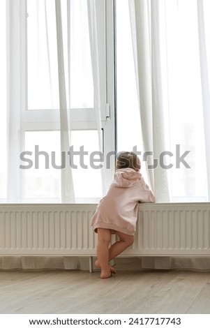Little child girl standing at window and looking outside. Back view. Royalty-Free Stock Photo #2417717743