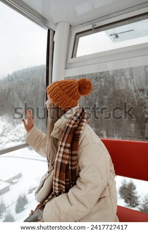 beautiful young girl dressed in winter outfit sits inside cabin of ferris wheel, snowy mountains and Christmas trees in background, winter entertainment at ski resort