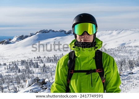 Close-up portrait of a skier or snowboarder in sports equipment, snowy mountains background at ski resort. Bright acid green outfit: warm suit jacket, goggles, black sport helmet, backpack straps Royalty-Free Stock Photo #2417749591