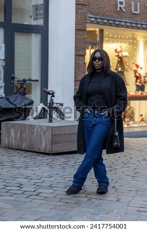 This image features a stylish young woman standing confidently on a cobblestone street. She is dressed in a black turtleneck and wide-leg denim jeans, paired with a long black coat. Her black ankle