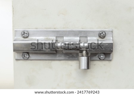 The barrel bolt or door knob or bolt made of stainless steel, which is a rust-resistant and durable material. It has a rectangular shape with rounded corners on white door,