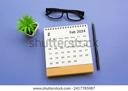February 2024 desk calendar with potted plant, pen and reading glasses on blue background. Royalty-Free Stock Photo #2417785087