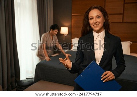 Cheerful housekeeping manager inspecting maid performance in hotel room Royalty-Free Stock Photo #2417794717