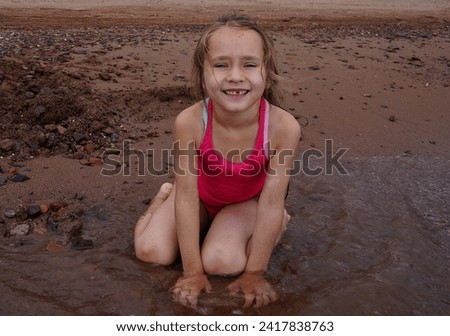 Little toothless smiling girl sitting on the seashore and playing in the sand. Traveling with children. Royalty-Free Stock Photo #2417838763