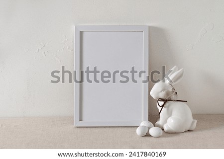 Minimalist white Easter holiday poster template, blank picture frame mockup, rabbit figurine and small candy eggs on neutral beige linen tablecloth background, textured empty concrete wall.