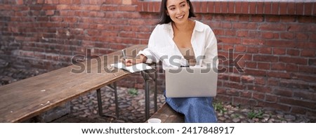 Working woman sitting outdoors in cafe, looking at laptop and making notes in notebook, writing down while watching smth on computer.