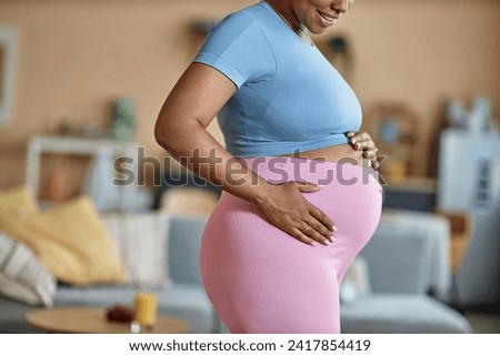 Side view cropped shot of smiling African American expectant mother in fitness outfit holding hands on her big belly