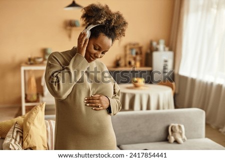 Medium shot of smiling African American expectant mother in headphones listening to calming music stroking round belly at home