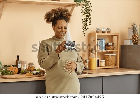 Medium shot of smiling pregnant Black woman holding ultrasound image of her baby while standing with hand on belly at kitchen counter with fresh vegetables