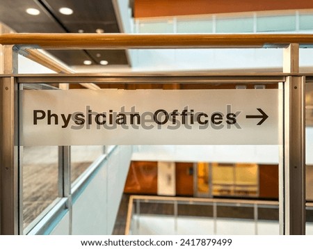 Transparent signage with arrow directing visitors to Physician Offices. Sign affixed to a balcony overlooking a modern office scene.