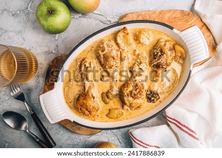 Chicken Normandy served in an enamel-coated cast-iron roaster, apples, glass of cider on a grey marble background. French cuisine. Fall recipe idea