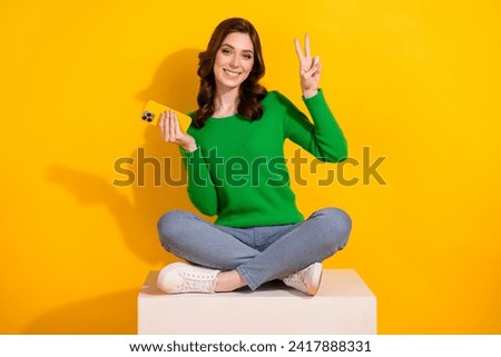 Full body length photo of young brunette woman promoting dating app with messenger use phone show v sign isolated on yellow color background