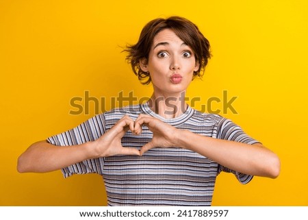 Photo of funny funky woman dressed striped t-shirt showing heart on chest plumb lips send kiss isolated on yellow color background