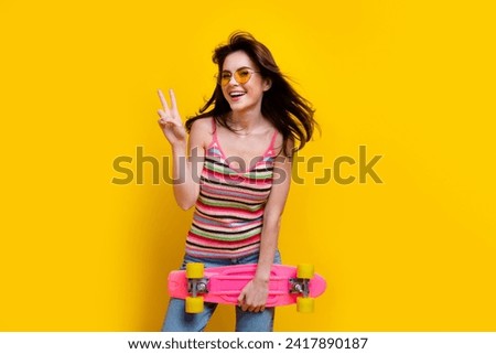 Portrait of good mood girl fluttering hair wear knit tank in glasses hold skateboard showing v-sign isolated on yellow color background